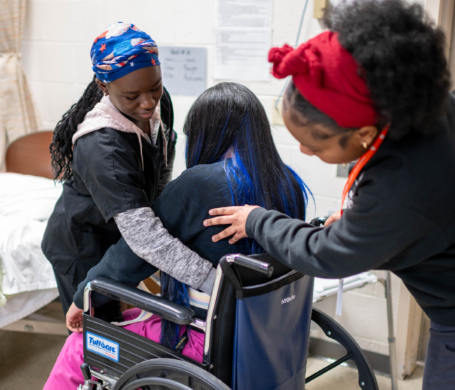 students assist a "patient" in a wheelchair